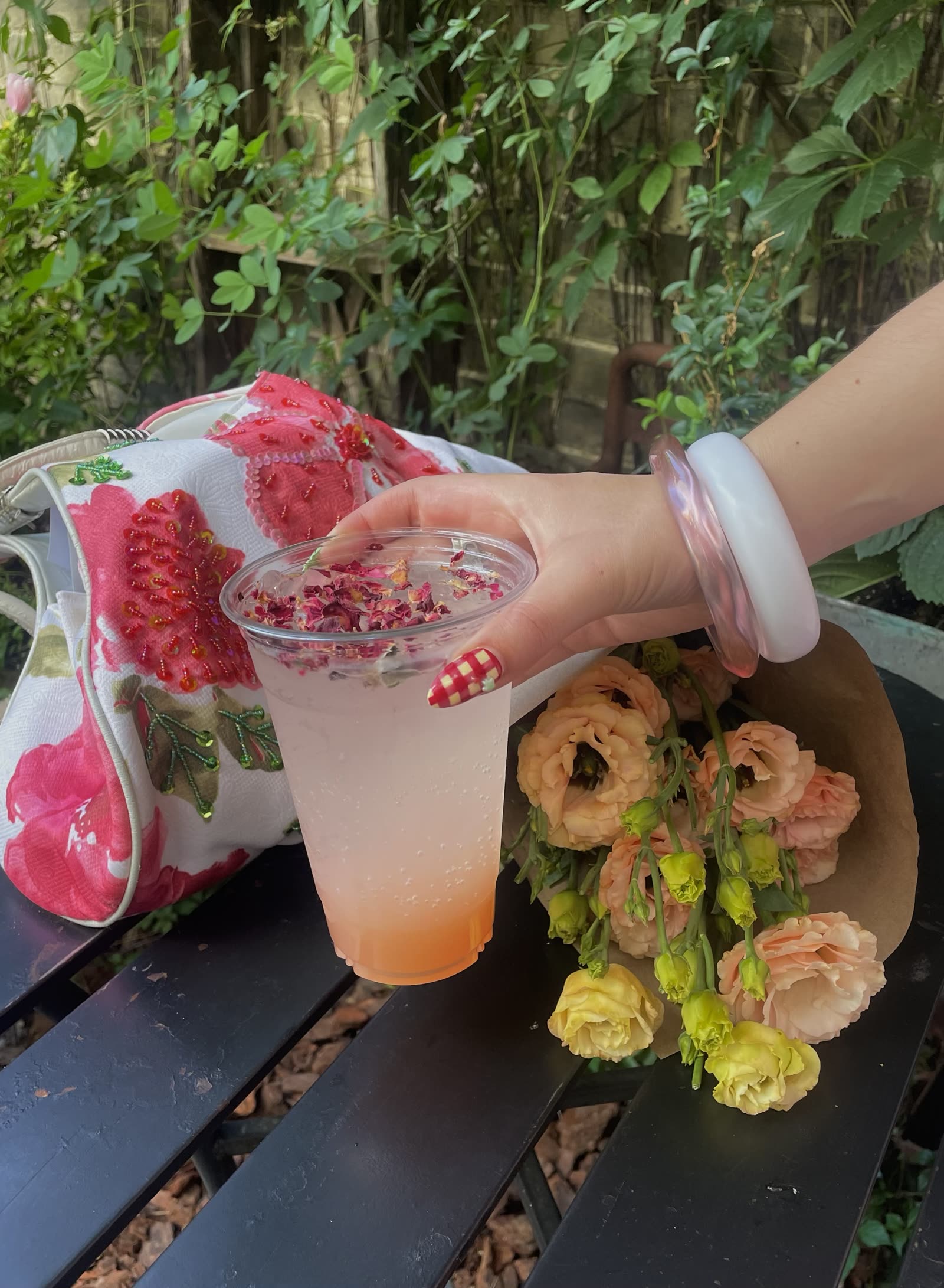 A gingham accent nail on a hand holding an iced rose drink, beside flowers on an outdoor table